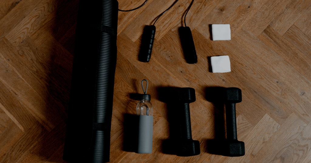 Black workout dumb bells, yoga mat, water bottle, and jump rope on a brown wood floor